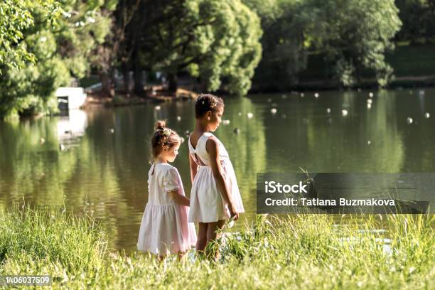 Cute caucasian and African-American girls in summer dresses watching birds on a pond on a summer day in the park,side view.Summer,diversity concept.