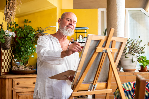 A confident and happy senior man enjoys his free time and life while painting a picture in his living room.