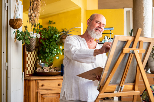 A confident and happy senior man enjoys his free time and life while painting a picture in his living room.