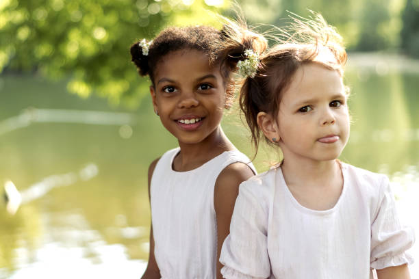 Happy caucasian and African-American girls in a summer dresses sitting on the shore of a lake in a park on a summer day.Summer,diversity concept