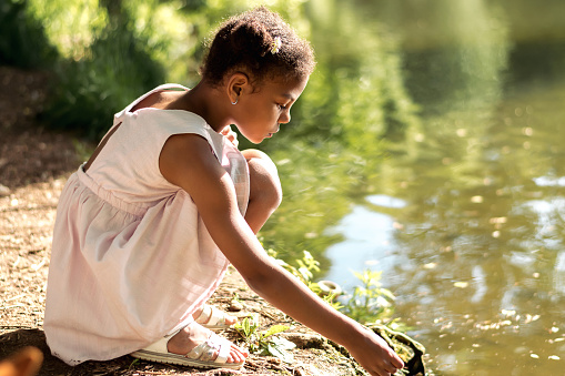 A cute African-American girl in a summer dress feeds birds on a lake on a summer day.Summer,diversity concept.Selective focus,copy space.