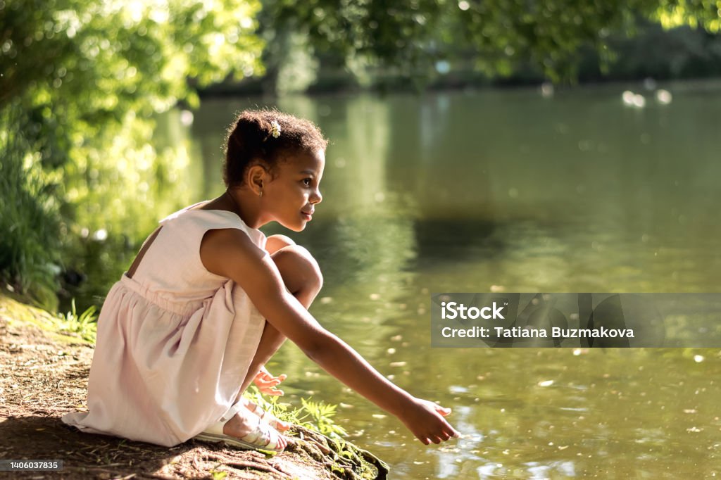 A cute African-American girl in a summer dress feeds birds on a lake on a summer day.Summer,diversity concept. A cute African-American girl in a summer dress feeds birds on a lake on a summer day.Summer,diversity concept.Selective focus,copy space. Child Stock Photo