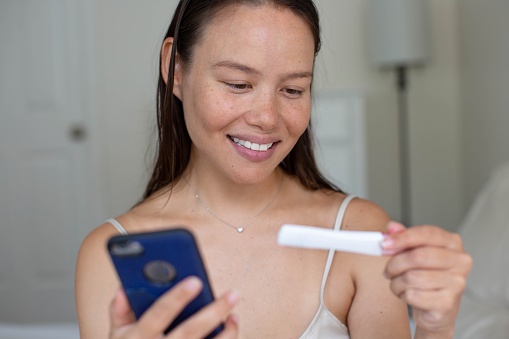A happy pleased woman smiling and holding a positvie pregnancy test and her phone