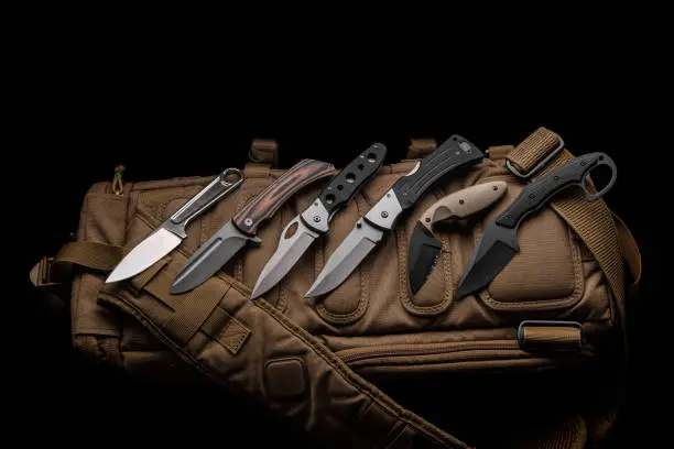 A variety of knives on a sand-colored military backpack. Dark background.