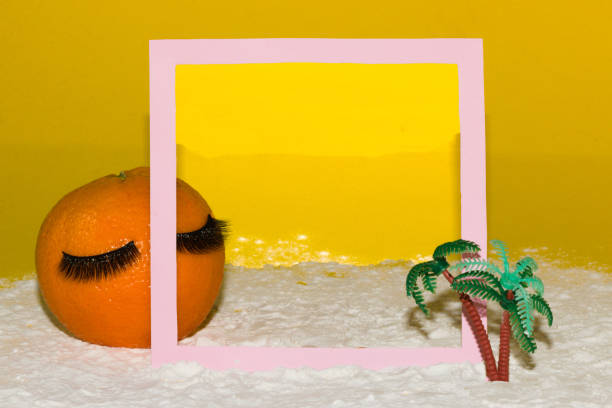 orange with eyelashes behind pink frame in which there is a sunset on the beach frame and orange are in the beach where there is a sunset, palm tree in front of the frame, relax tropical