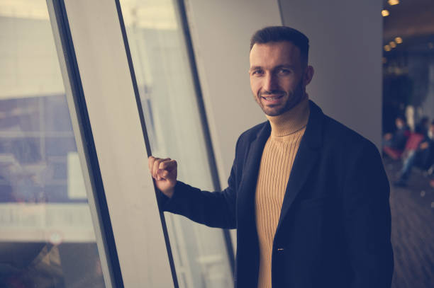 istock Handsome young man, transit passenger, traveler, smiling at camera, standing near panoramic windows overlooking runway in airport departure terminal 1406051996