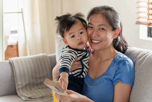Happy Asian Mother embracing her baby son and holding wad of 1,000 Thai Baht banknotes, smiling and looking at camera.