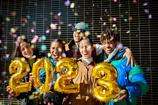 Waist-up view of diverse men and women in teens and 20s standing together, holding gold foil number balloons, and grinning in Barcelona sunshine.