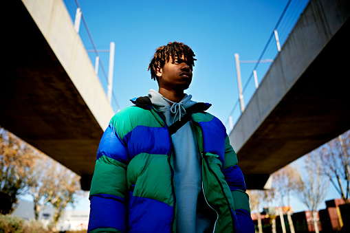 Waist-up view with diminishing perspective of man wearing ski jacket over hooded sweatshirt, standing beneath elevated roads, and looking away from camera.