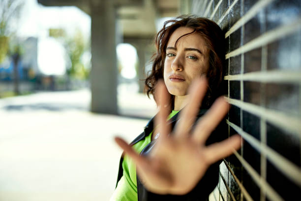Waist-up side view of brunette woman in mid 20s standing outdoors in urban area, leaning against wall, and holding up palm to camera.