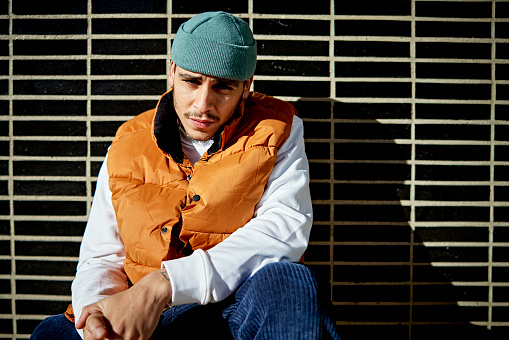 Front view of man in winter clothing sitting outdoors against black wall with geometric pattern and staring at camera with negative expression.