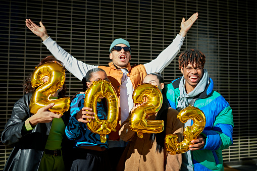 Waist-up view of multiracial group in teens and 20s standing beneath man with arms outstretched, holding gold foil number balloons, and smiling.