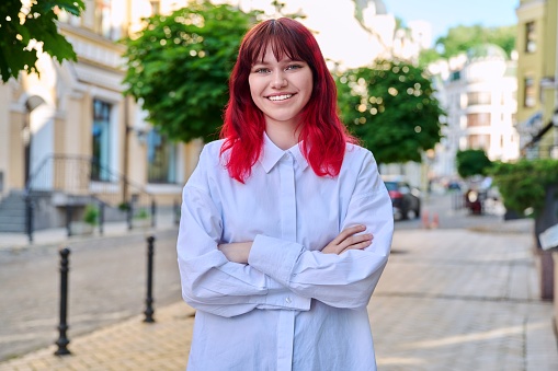 Smiling confident young female with crossed arms looking at camera. Teenage beautiful fashionable girl with red dyed hair in city. Beauty, youth, lifestyle concept