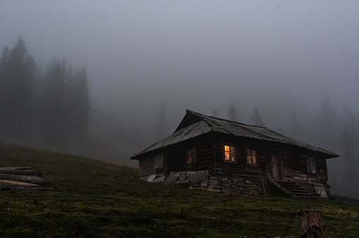Lonely hut in the forest. Lonely wooden house in the mountains. Gloomy house with luminous windows.