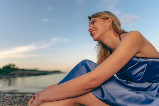 Young blonde-haired woman in a blue dress is sitting by the sea at sunset.