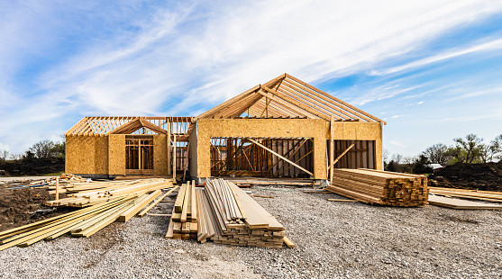 New house under construction, front view from garage door entry with clouds blue sky. Framing of home exterior structure with a pile of lumber and siding plank leftover. Real estate business industry.