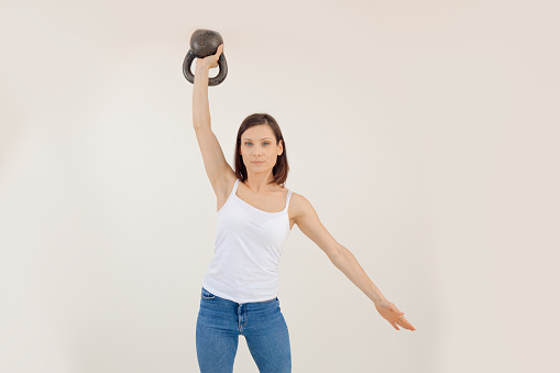 Young athletic woman lifting up kettlebell and putting down arm, white background. Keeping fit by strength workout and weightlifting in gym, pumping up muscles. Sport and healthy lifestyle concept.