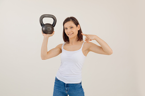 Young athletic woman lifting up kettlebell and enjoying training, white background. Keeping fit by strength workout and weightlifting in gym, pumping up muscles. Sport and healthy lifestyle concept.