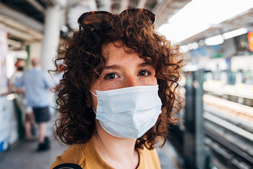 A portrait of a Caucasian woman with curly ginger hair, standing on a railway platform wearing a face mask.