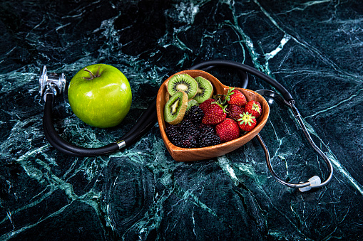 This is an overhead photo of a heart bowl of fruit and an apple on a green marble background with a doctor stethoscope
