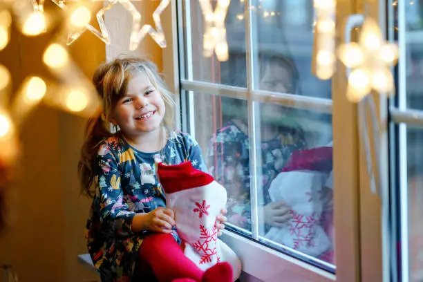 Little preschool girl holding cup Santa Claus boot with gift called Nikolausstiefel in German. Happy child wait on holiday by window with Christmas lights in winter. Cozy family celebration of xmas