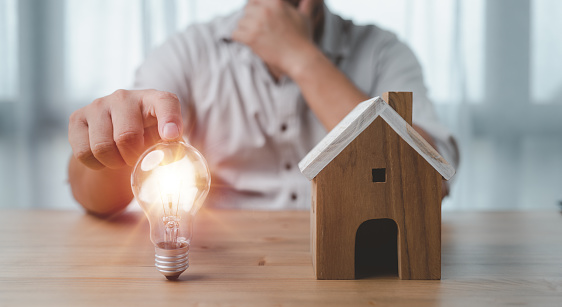 male hand touching a light bulb with house model on desk. The concept of installment and reduction of home loan interest, saving energy and money concept. idea for Buying a house.