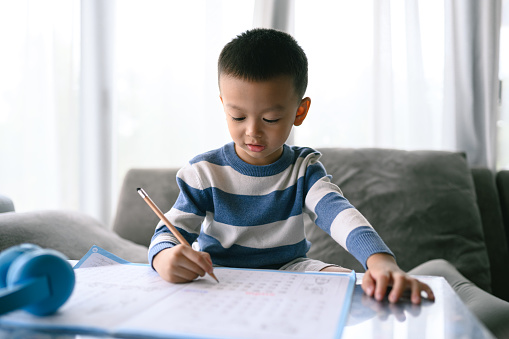 Asian Boy Doing homework with the intention. Child boy holding pencil writing, A boy drawing on white paper at the table, Elementary school and home schooling, Distance online Education concept.