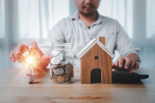 The concept of installment and reduction of Home Loan, Interest Calculation, Taxation, saving energy and money concept. male hand touch a light bulb with coins stacking and house model on desk.