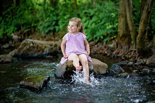 Cute little toddler girl having fun by a river on warm and sunny summer day. Happy excited preschool child splashing with water in forest stream creek