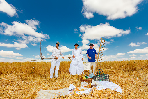 Muzlja, Vojvodina, Serbia, - July 03, 2021; XXXVIII Traditionally wheat harvest. Cheerful farmers prepare for mowing wheat, hold old harvest tools, scythe and wooden rake. Breakfast in retro style.