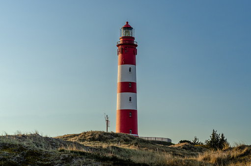 Red and white striped lighthouse against a clear evening sky in the dunes. The dunes have light vegetation.