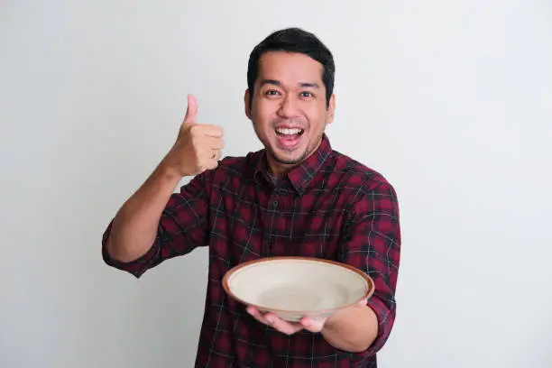 Adult Asian man give thumb up and showing enthusiastic expression while holding empty dinner plate