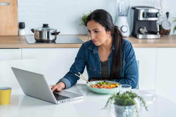 istock Cheerfull woman working with computer while with computer while eating poke bowl in the kitchen at home. 1406144970