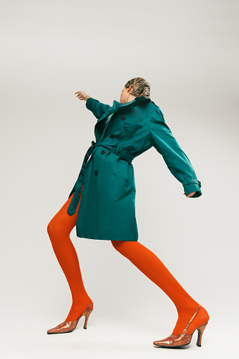 Side view portrait of stylish girl in green coat and bright orange tights dancing isolated over grey background. Expressive fashion. Concept of retro fashion, art photography, style, queer, beauty
