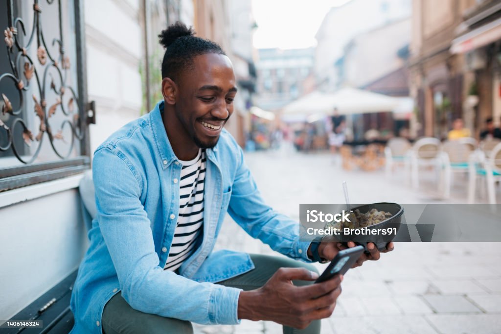 Eating a take out food outdoors Happy young adult man sitting on a wooden bench outdoors in the city, eating and enjoying a take out meal while using a smart phone Eating Stock Photo