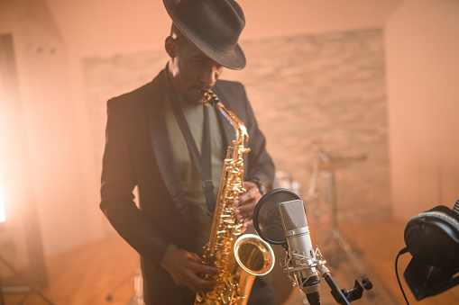 Black mid-adult male musician playing saxophone and recording songs in recording studio. Waist-up, looking away.