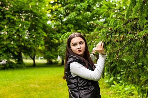 Portrait teenage girl walking in spring park in green fir trees or spruce. Happy teen in casual clothes at summer natural park. Caucasian student girl lifestyle. Countryside concept. Copy space