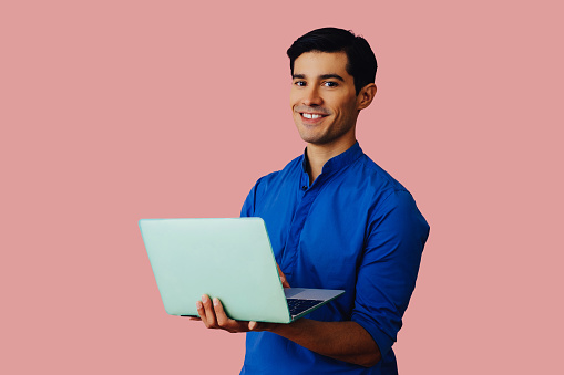 Portrait hispanic latino entrepreneur man holding laptop black hair smiling handsome young adult blue shirt over pink background looking at camera studio shot