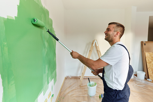 Happy male worker painting the wall in green color during home renovation process. Copy space.