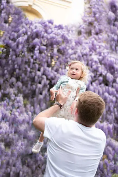 Father play,having fun outdoor with baby girl,daughter on background of glucinum flower,purple,very peri wistaria tree.Man and toddler laughing smiling.Happy parenthood,childhood idea, lifestyle.