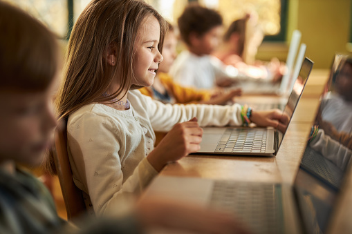 Happy schoolgirl and her classmates using computers for e-learning on a class at elementary school.