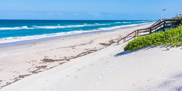 White sand beach in Florida. In the distance, the waves of the ocean, a wooden staircase to the beach and a weather vane with the symbols of the American flag
