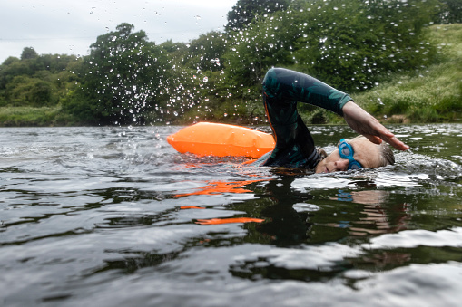 A side-view shot of a mature woman swimming in a lake during the day in the North East of England. She is using a swimming float and looking forward.