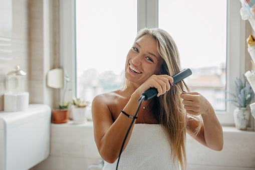 A young Caucasian woman is wearing a bath towel and straightening her hair, while looking at the camera with a smile on her face.