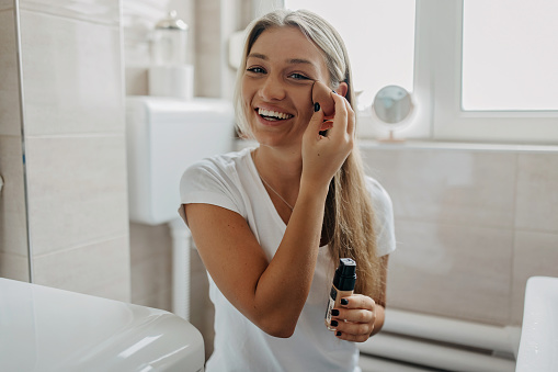 A young Caucasian woman is doing her make-up with a big smile on her face.