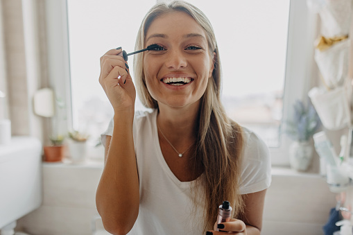 A young Caucasian woman is smiling wide while putting on her mascara and looking at the camera.