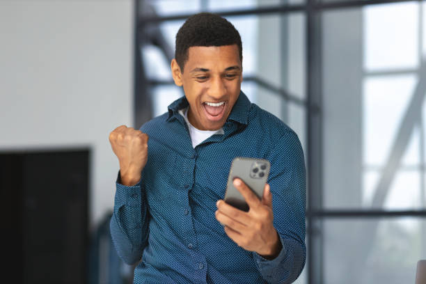 Excited African American man holding a mobile phone, showing yes gesture, feeling euphoric about good news, he receive payment. Young male businessman delighted with receiving mail on smartphone stock photo