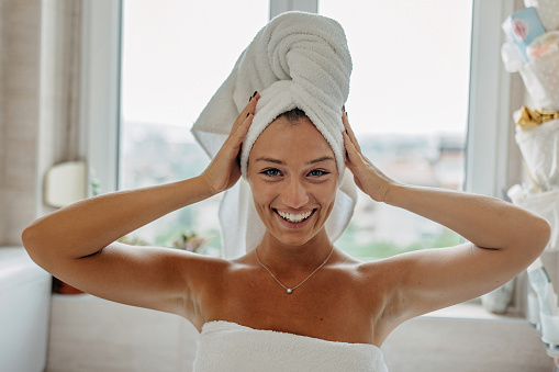 A young Caucasian woman is looking at the camera, while holding her arms up to her hair that she wraped in a bathroom towel.