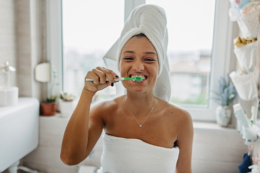 A young Caucasian woman is wearing a towel on her hair and looking at the camera while brushing her teeth.