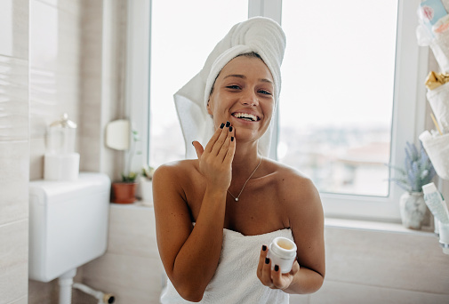 A young Caucasian woman is looking at the camera with a big smile on her face, while applying a face cream and wearing her hair up in a towel.